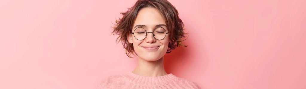 Waist up shot of relaxed happy young woman makes zen gesture meditates indoor breathes deeply practices yoga to feel relaxed wears spectacles and casual jumper isolated over pink background.